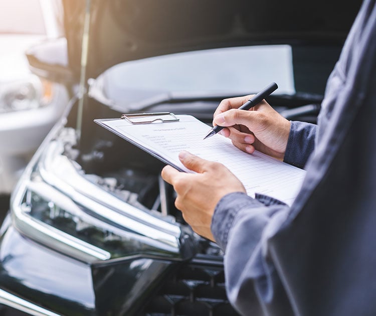 A mechanic with a clipboard under the hood of a car.