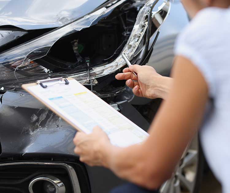 An insurance adjuster with clipboard reviews a damaged car.