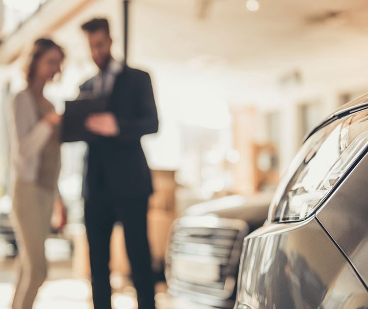 Car in the foreground with a blurred couple in the background reviewing a handheld document.