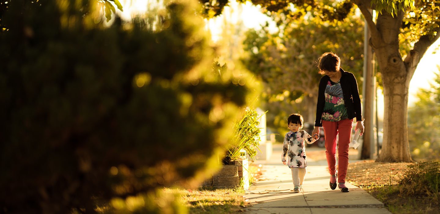 An adult and infant holding hands walking on sidewalk.