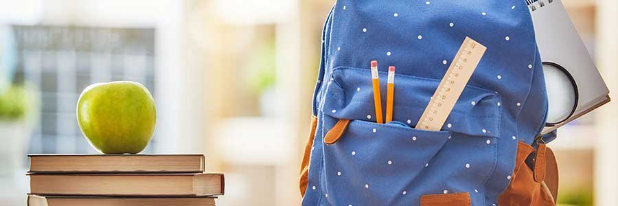 Image of an apple on school books next to a spotted blue kid's backpack.