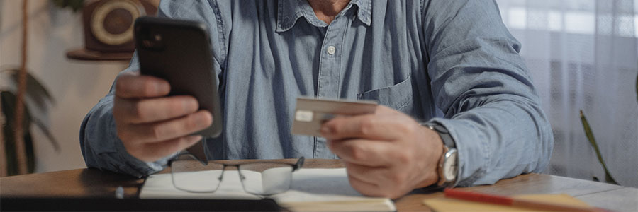 Photo of holding a mobile device and credit card with financial documents spread out on a kitchen table.