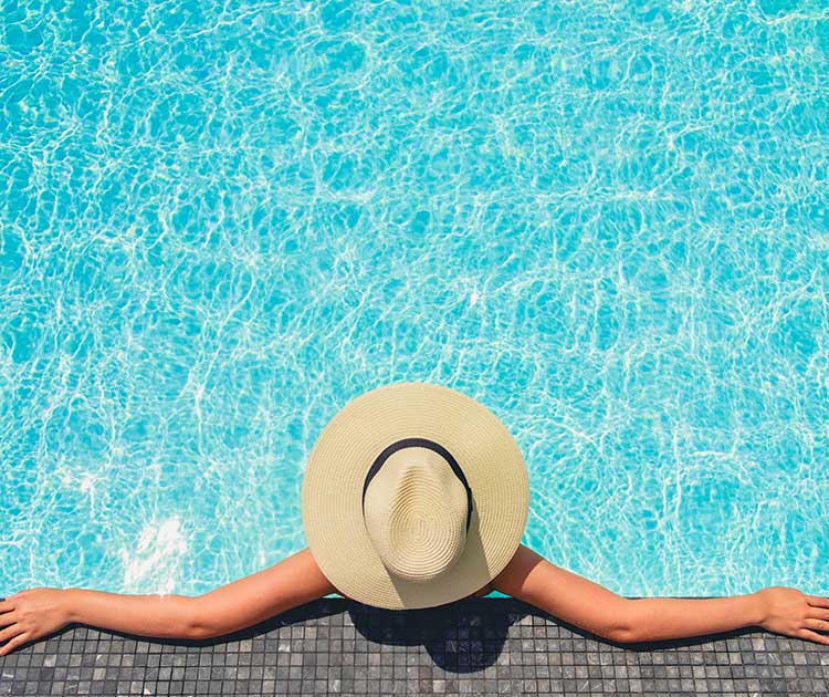 A top-down image of a woman with a straw hat relaxing in a pool with arms out.